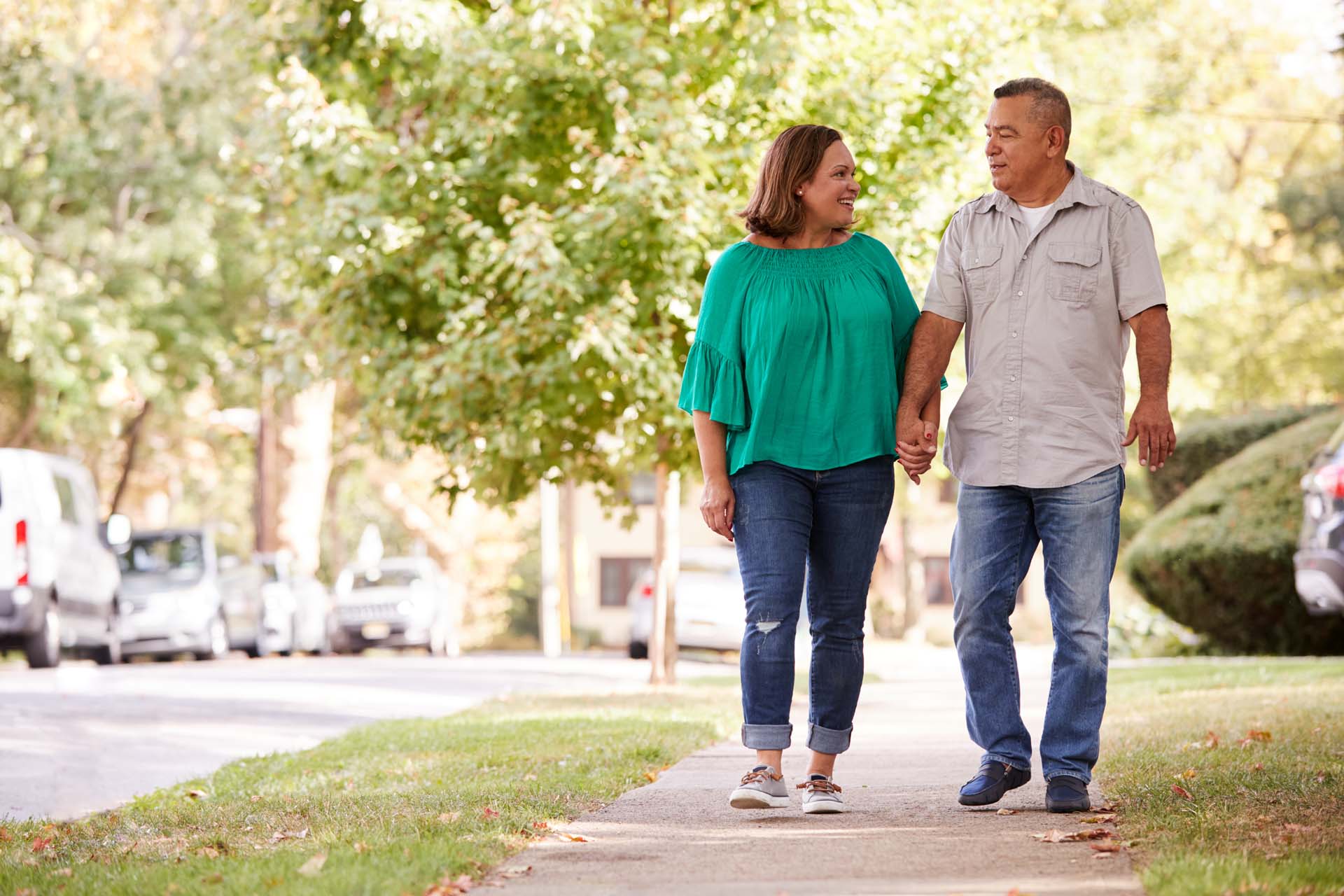 An older couple holding hands walking along a pavement next to a tree-lined road.
