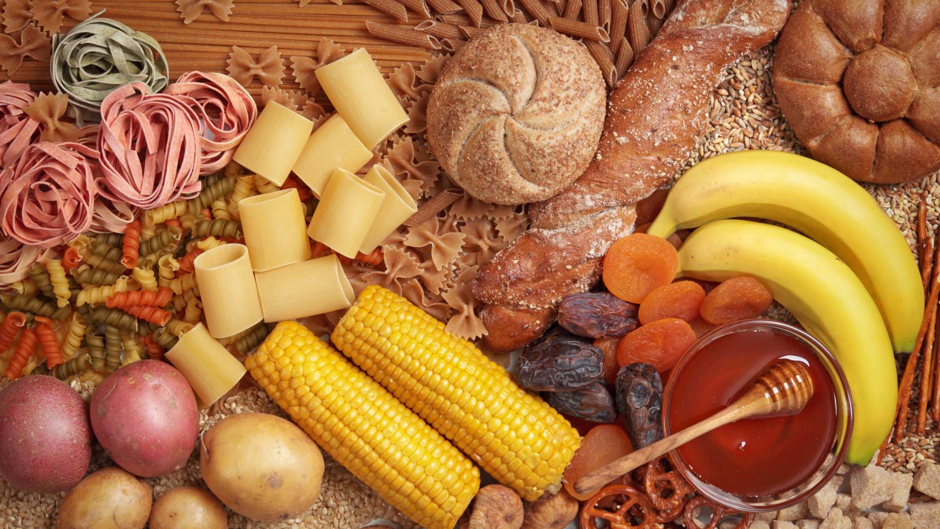 A selection of carbohydrates including pasta, bread, honey and sweetcorn laid out on a table.