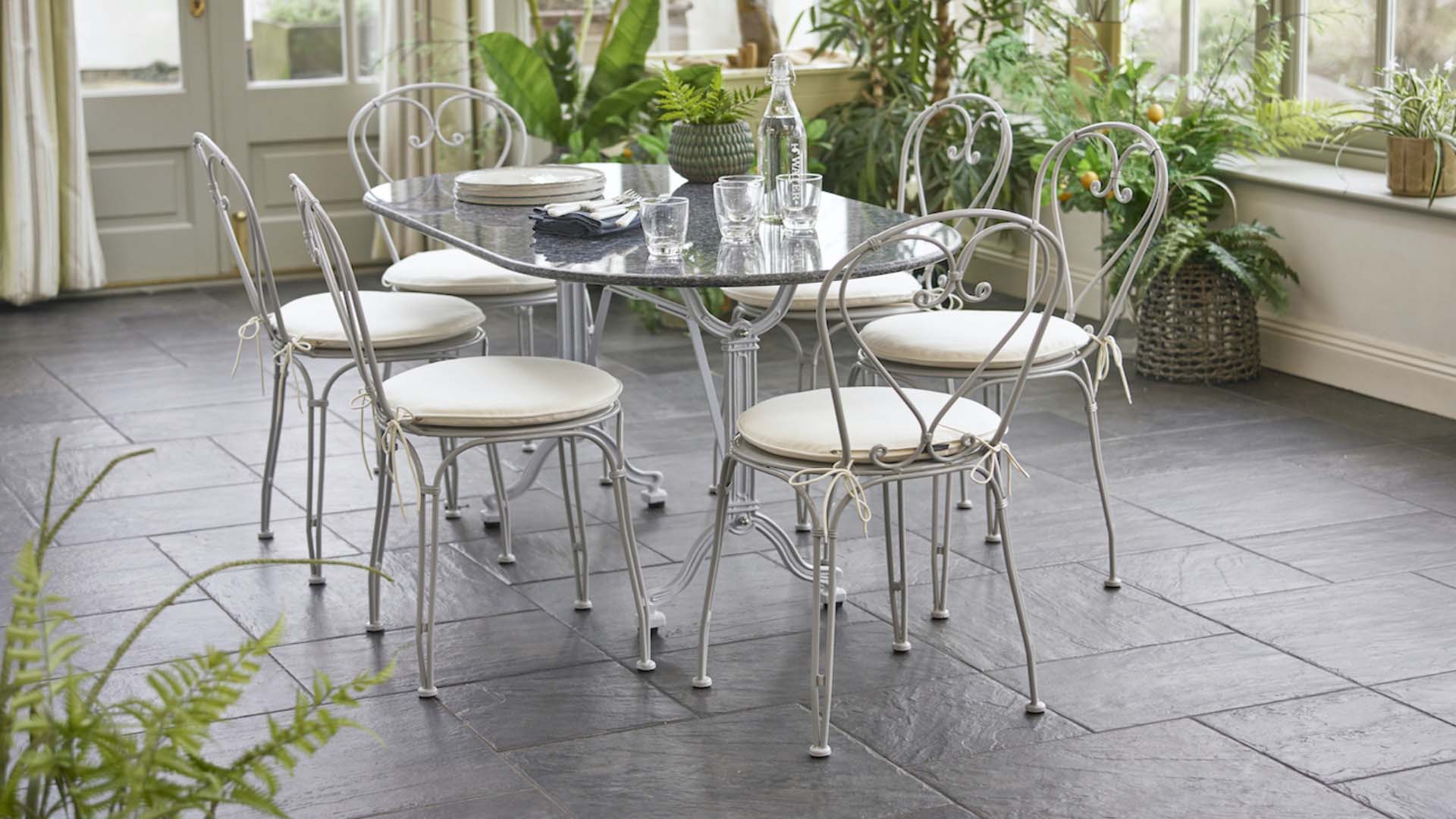 Traditional outdoor dining set with six white metal chairs and a glass-topped table, inside a conservatory.