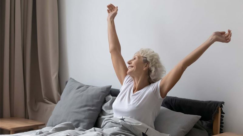 Optimistic refreshed middle aged senior woman sitting in bed wake up feel rested after enough healthy sleep in comfortable bed, stretching arms smiling looking out the window enjoy and welcome new day