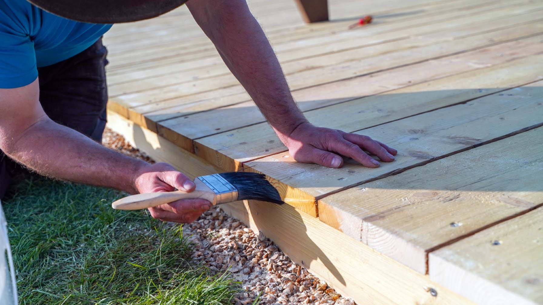 A man kneeling on the grass oiling the new wood deck