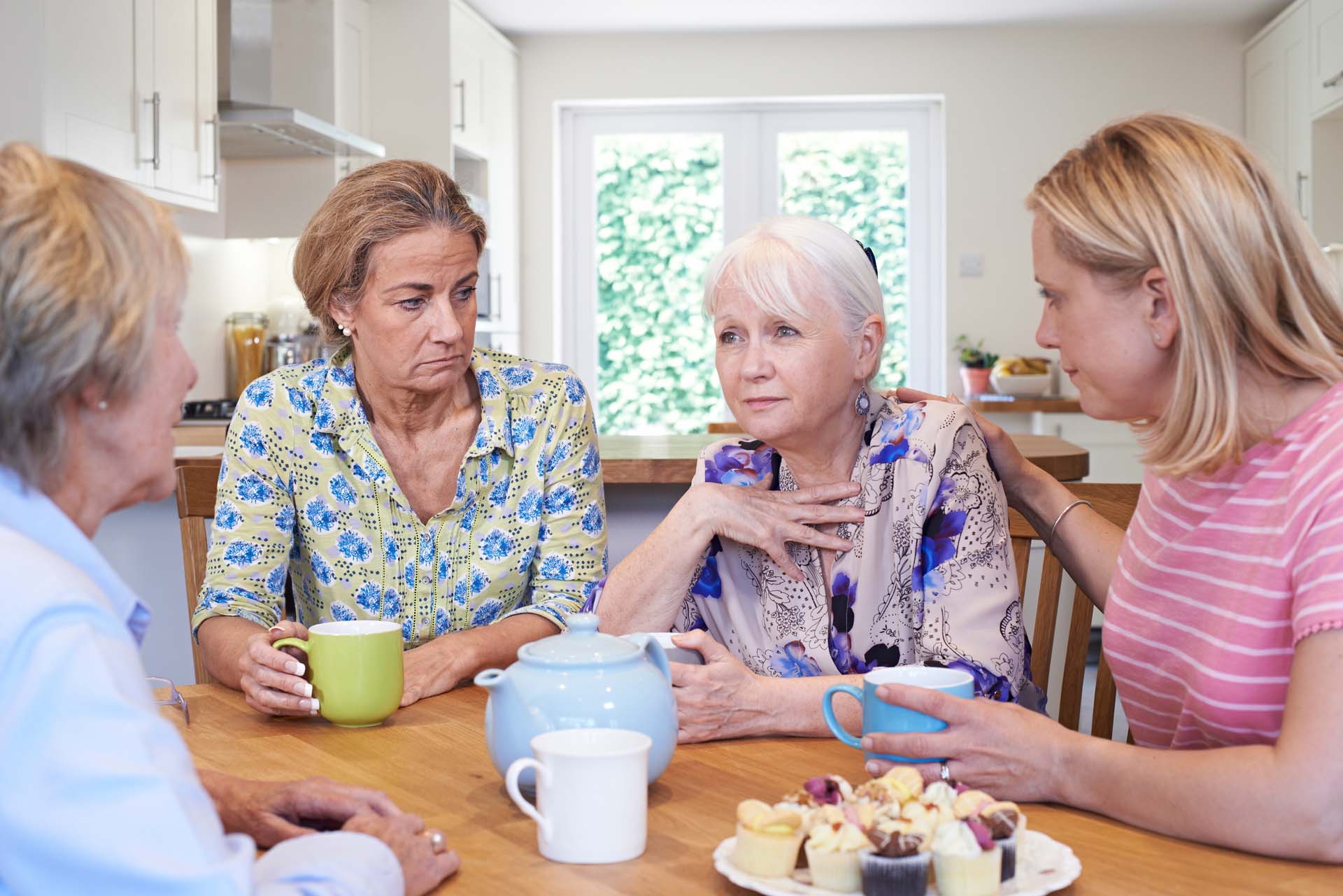 A group of women supporting an elderly friend around a kitchen table over a cup of tea