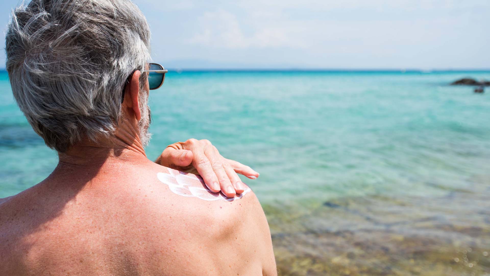 A man rubbing suncream into his back on a beach overlooking the sea