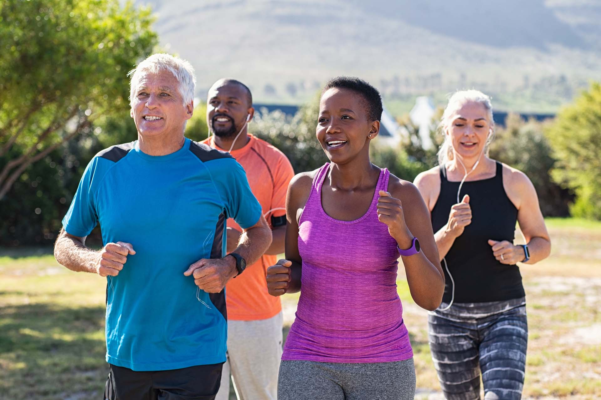 Four multiethnic middle-aged men and women running in sports attire on a sunny day in a park with trees.