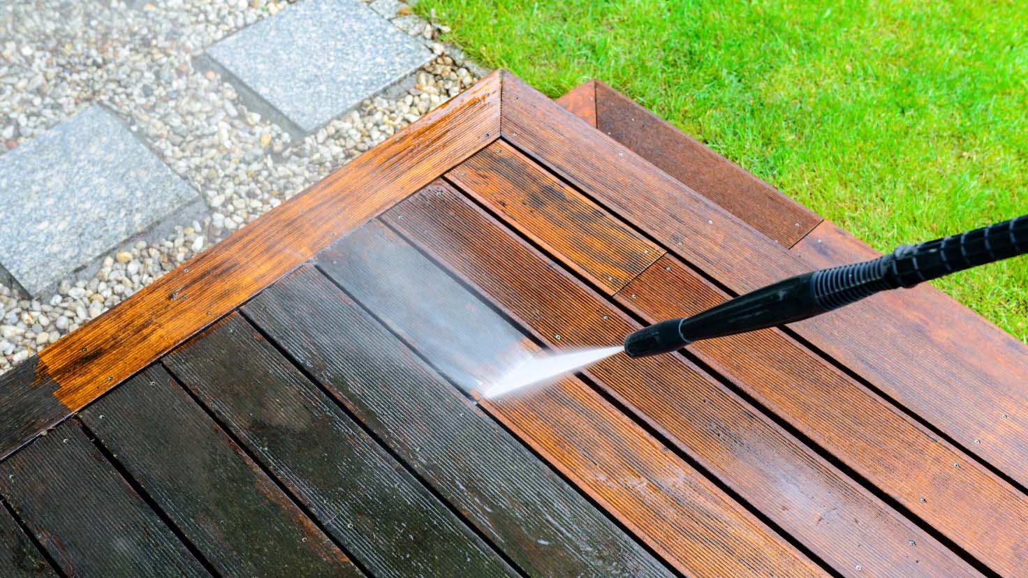 Someone cleaning a wood terrace sided by a grass area in the right and gravel  on the left, using a high water pressure cleaner