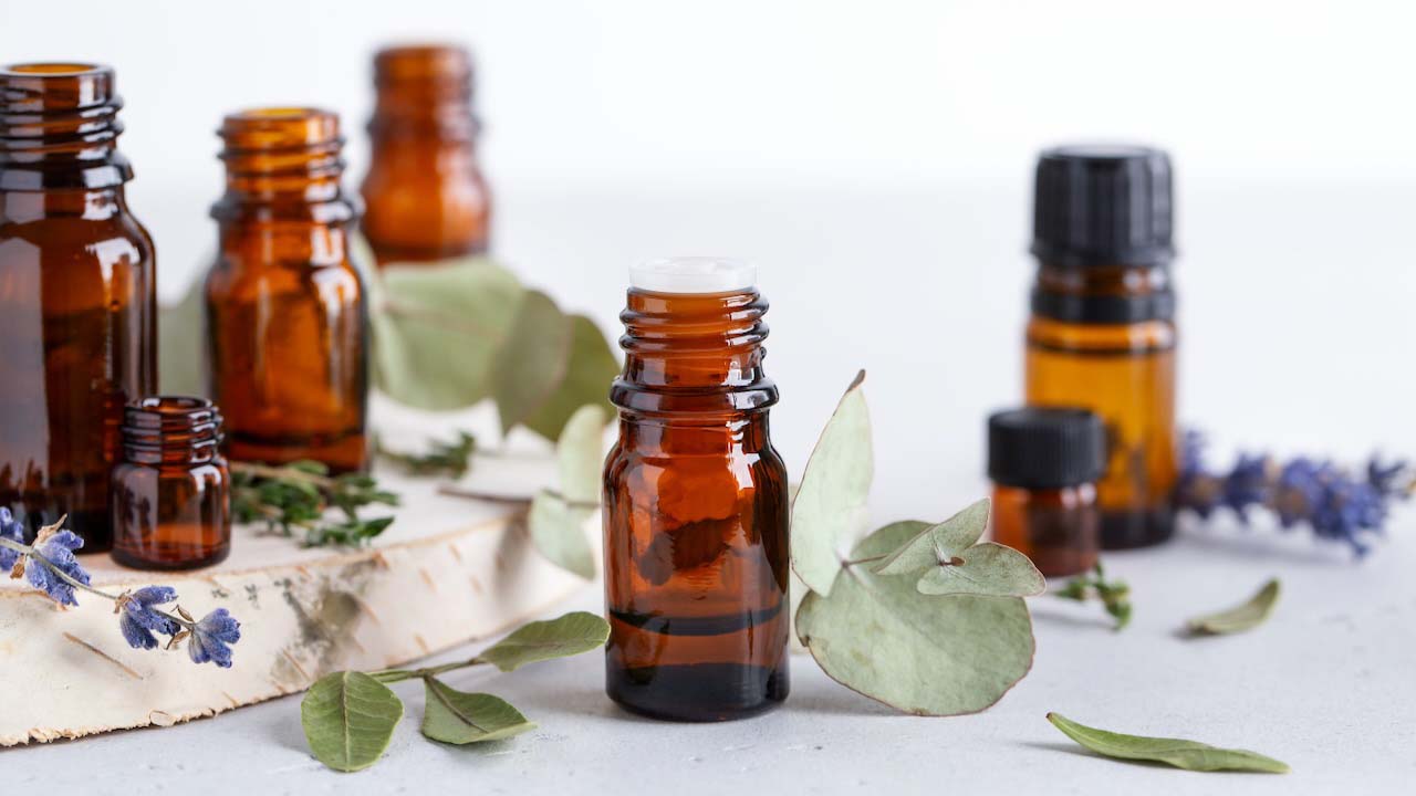 Collection of various essential oils in amber bottles with different herbal ingredients - mint, rosemary and eucalyptus leaves on neutral background