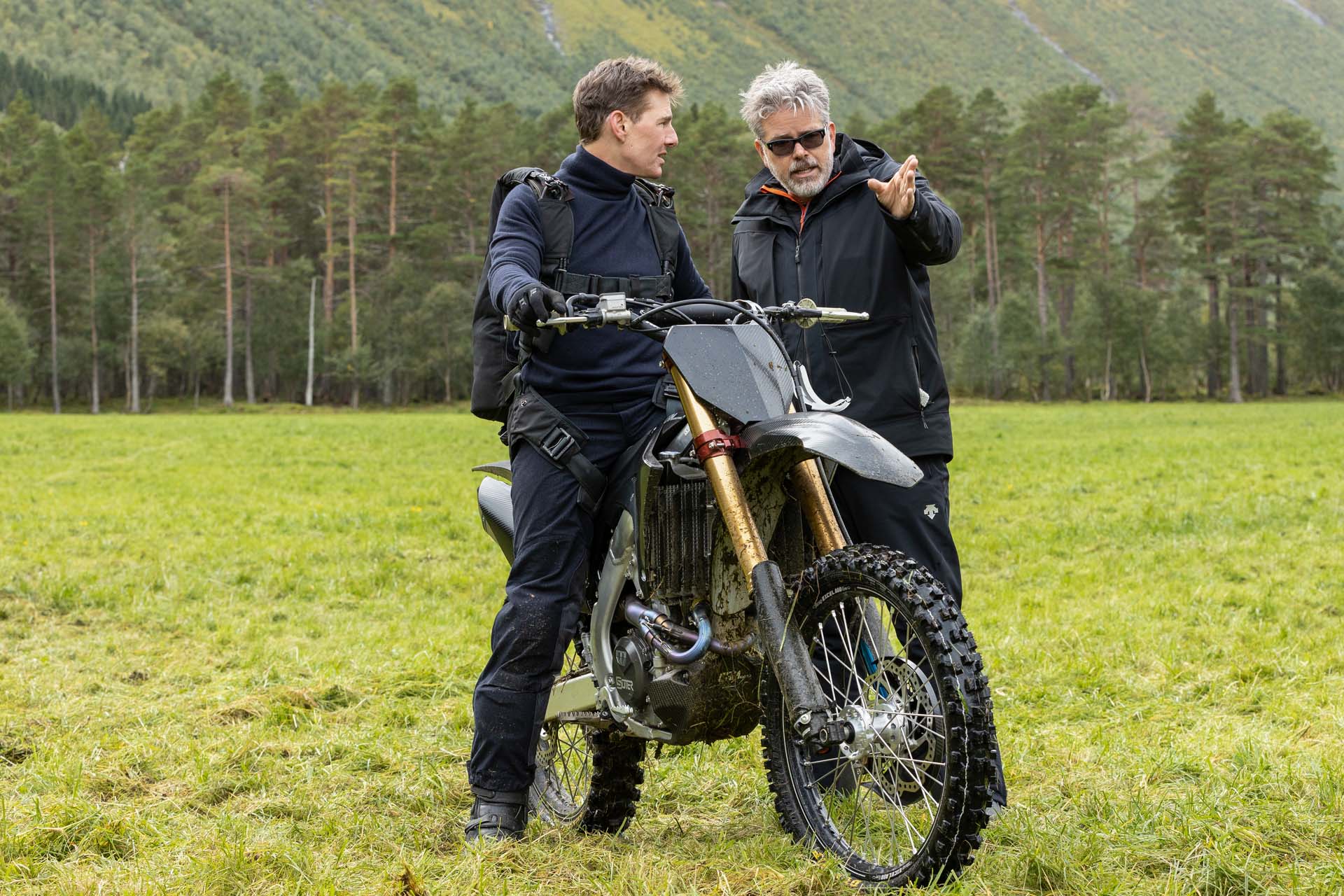 Actor Tom Cruise sitting on a motorbike in a large grassy clearing talking to film director Christopher McQuarrie on the set of Mission: Impossible Dead Reckoning