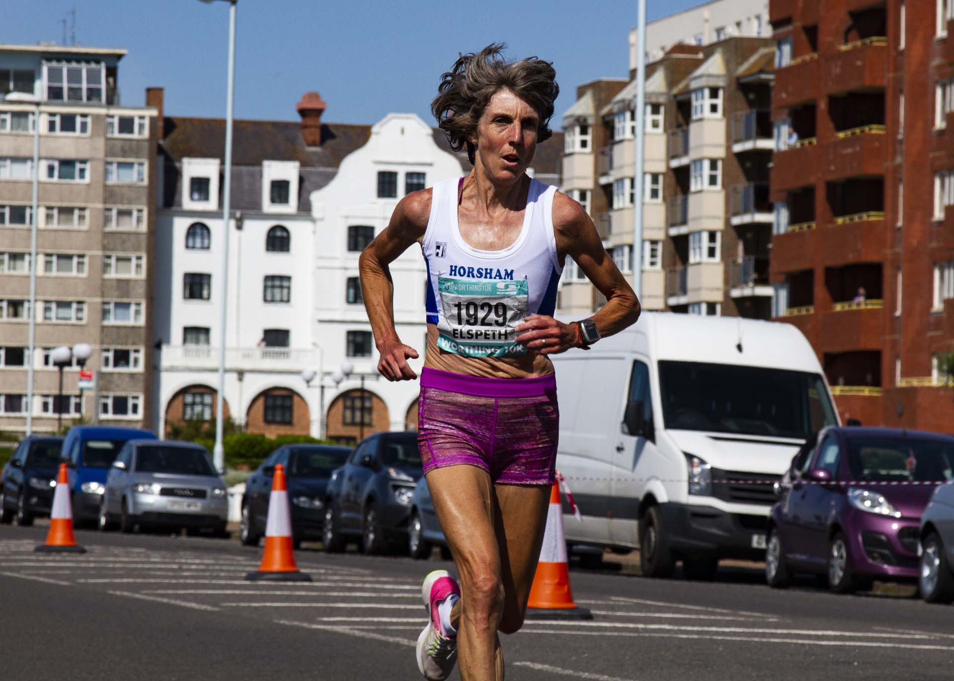 Elspeth Turner, a 58-year-old runner and coach, completing an organised run through a city wearing running attire and a number pinned to her front.