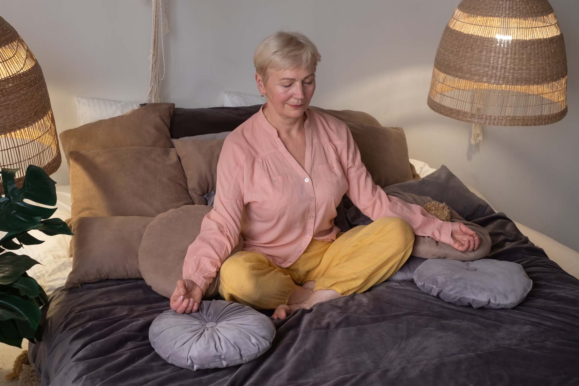 An older women sits cross-legged on a bed surrounded by cushions and lamps, with hands resting on her knees and eyes closed in a yogic meditation.