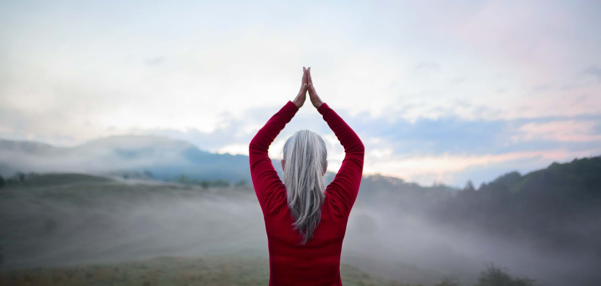 A woman looks out over a mountainous view while performing yoga with her hands raised and palms together above her head.