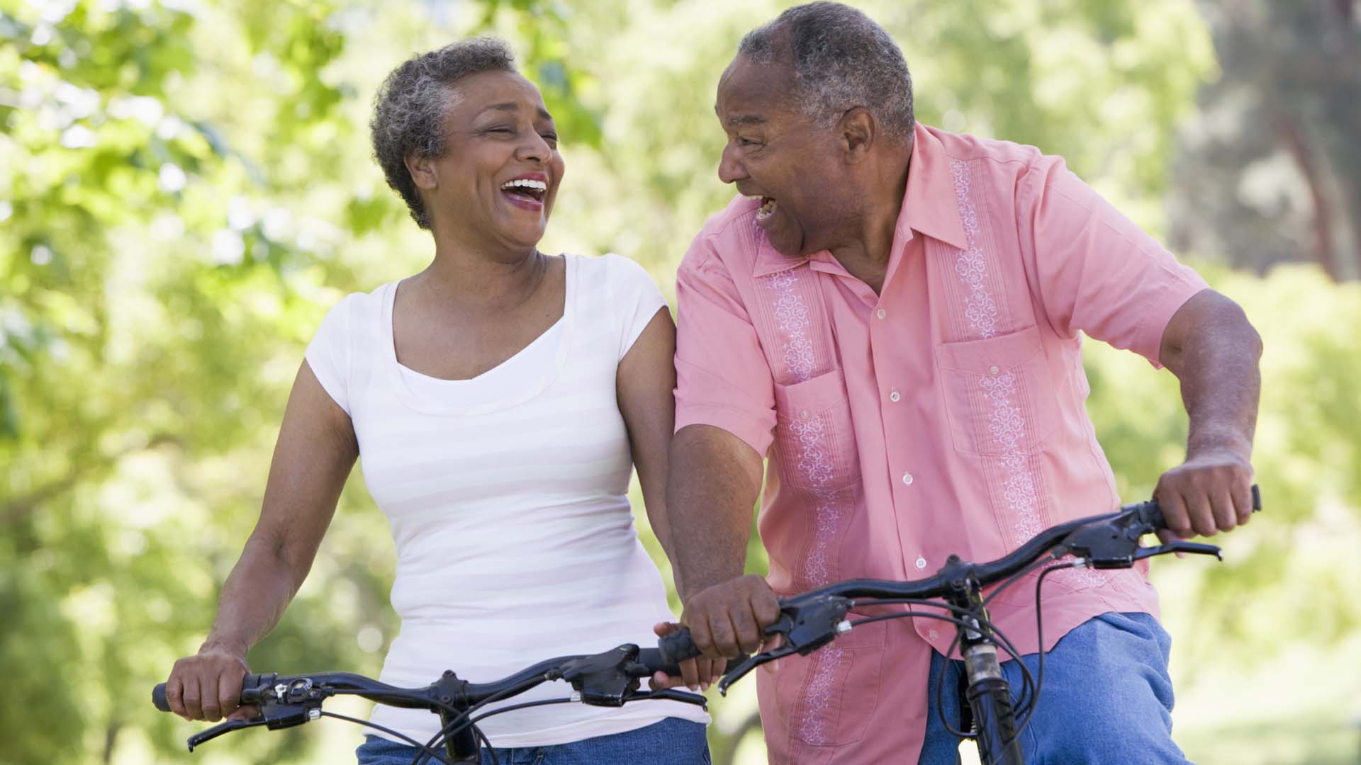 A close-up of two smiling adults riding bikes with foliage visible behind them