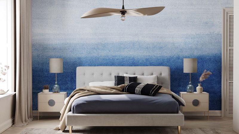 A bedroom with an ombre-effect wall in shades of blue and white, with a dhouble bed in shades of grey and bedside tables.