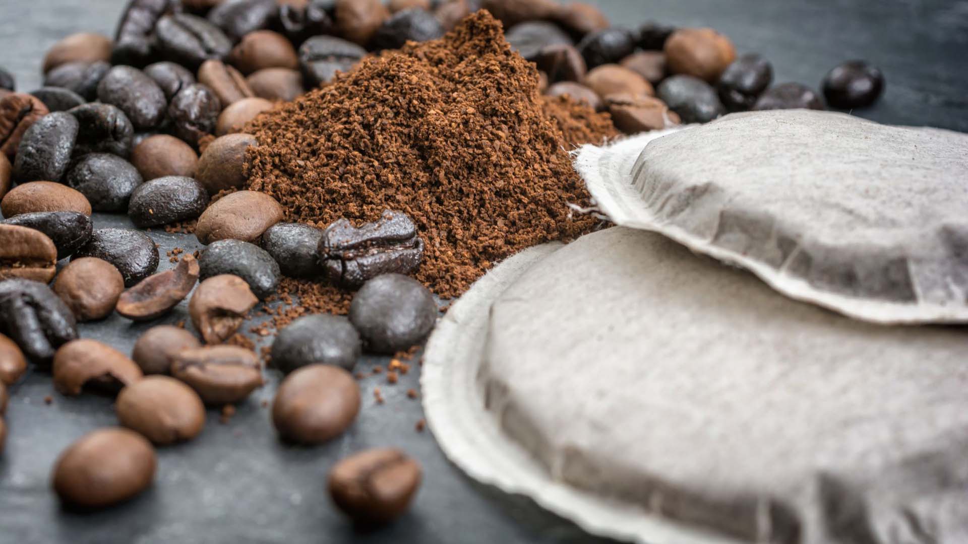 Close-up of coffee beans, coffee grounds and some tea bags on a dark grey surface.