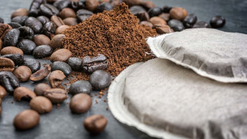 Close-up of coffee beans, coffee grounds and some tea bags on a dark grey surface.