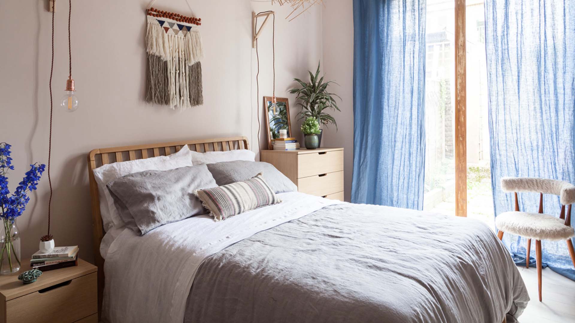 A bedroom with wooden bedside furniture, airy blue curtains and grey soft furnishings