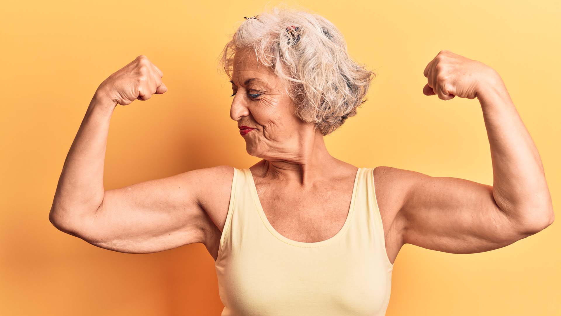 A woman in a yellow vest top flexing her arm muscles