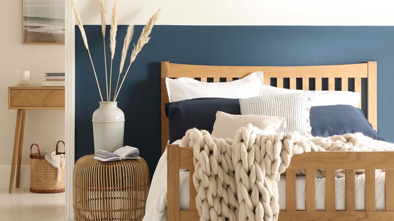 A bedroom in shades of blue and white with a wooden double bed frame covered in light bedding and a wicker bedside table.