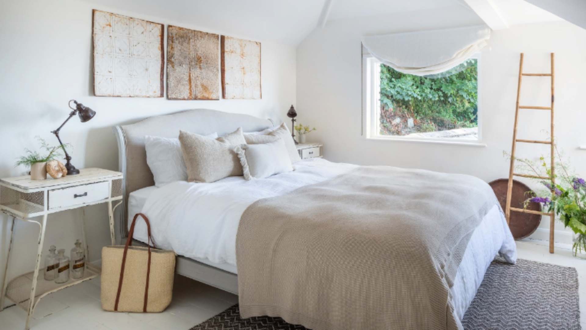 A light-coloured bedroom with double bed in soft grey colours, a desk-style bedside table topped with a lamp, art hanging above the bed and a decorative ladder propped beside a window.