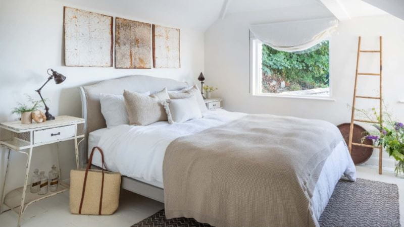 A light-coloured bedroom with double bed in soft grey colours, a desk-style bedside table topped with a lamp, art hanging above the bed and a decorative ladder propped beside a window.