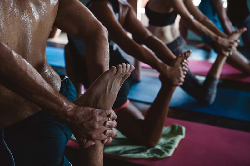 The back of a shirtless man and other people in sports attire practising hot yoga; they pose on one knee with their back foot stretch up behind them.