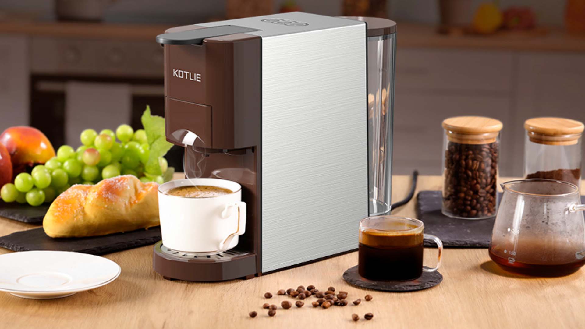 A coffee machine with a mug of espresso, surrounded by jars containing coffee and a selection of foods in the background, such as grapes, a croissant and an apple.