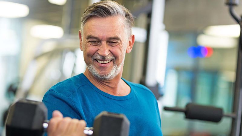 An older man with short grey hair and facial hair wearing a blue t-shirt lifts a hand weight in a gym environment.