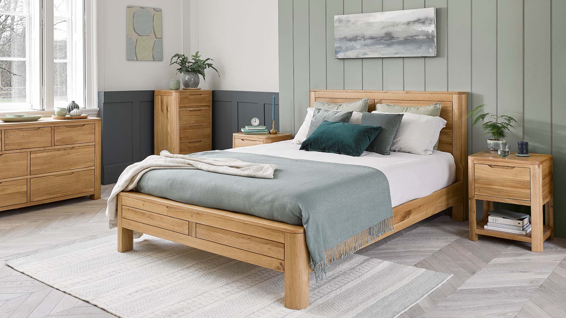 A bedroom decorated in grey-green and white shades containing a double bed, wooden furniture and coastal-inspired art