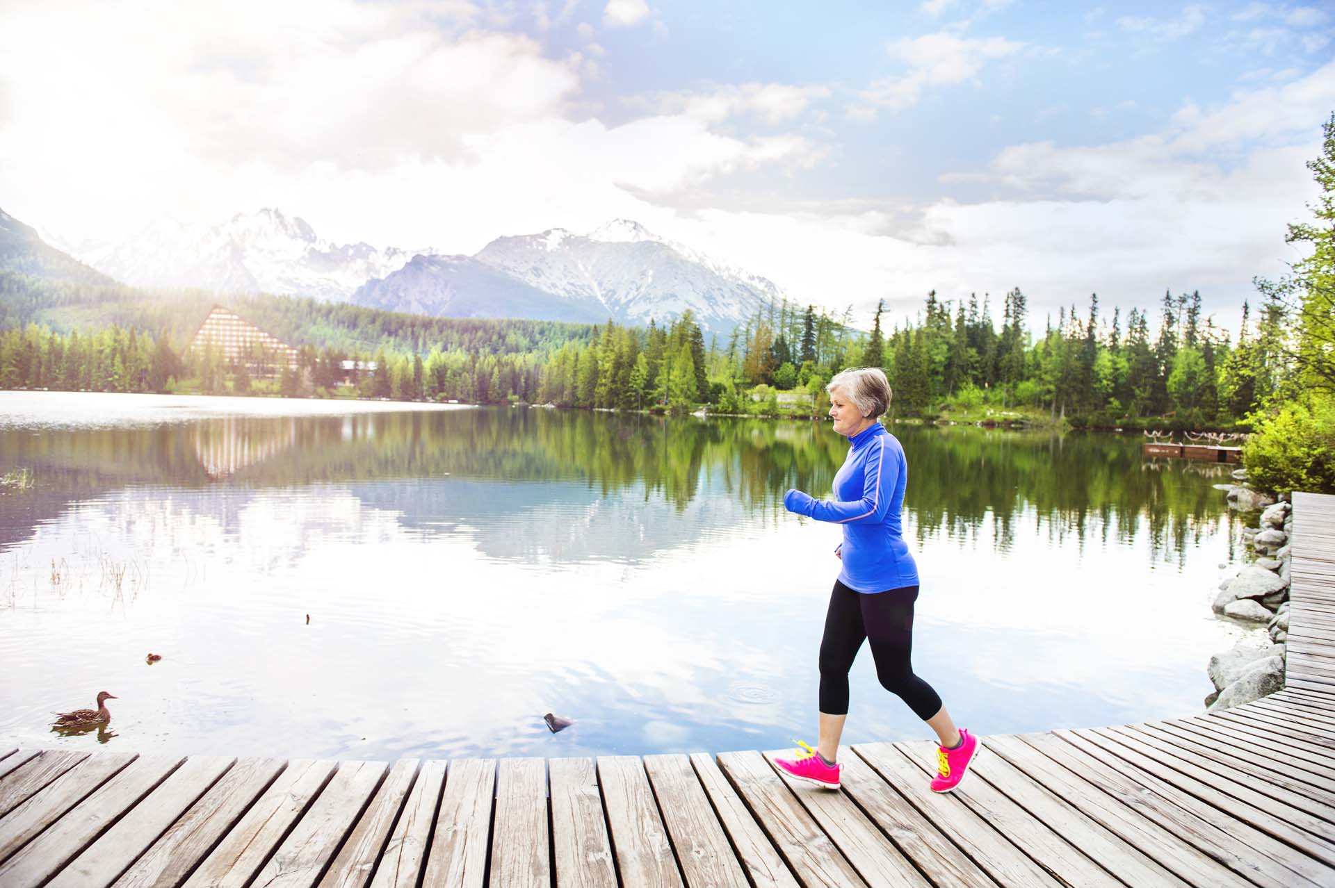 older woman running along a wooden path by a lake with mountains in the background