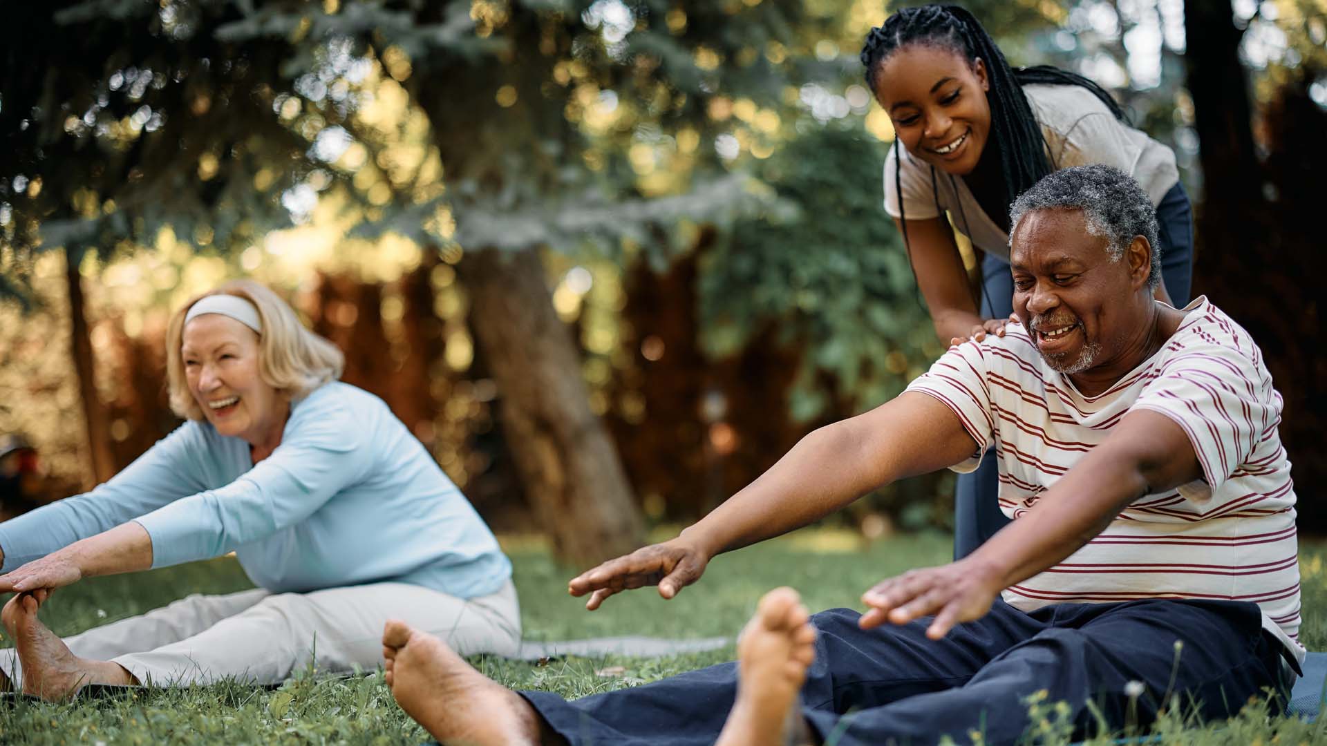 A black man and white women sitting in a grassy park stretching to reach their toes with another woman watching and trees visible in the background.