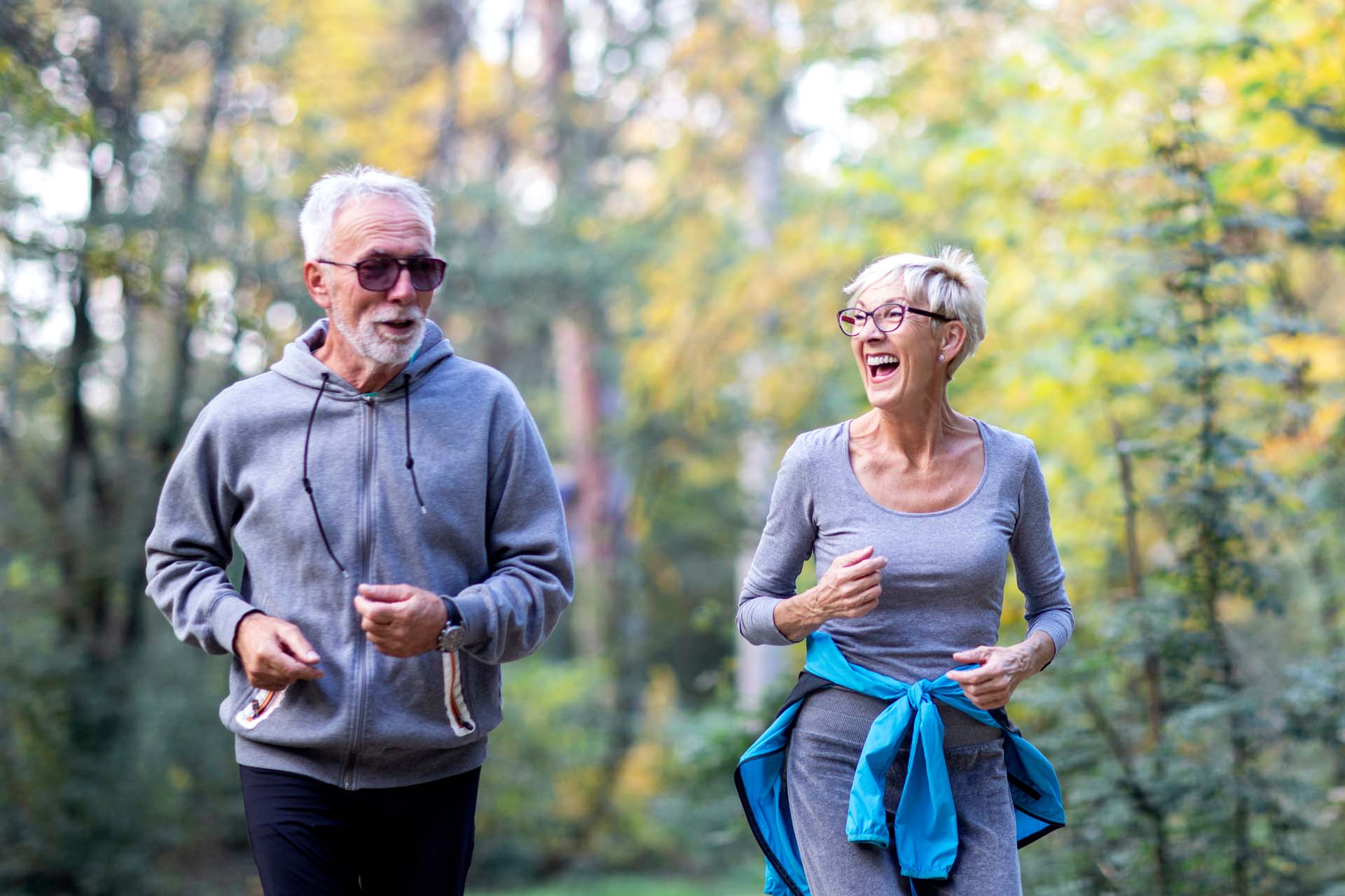 An older couple running outdoors in sports gear, with trees visible in the background