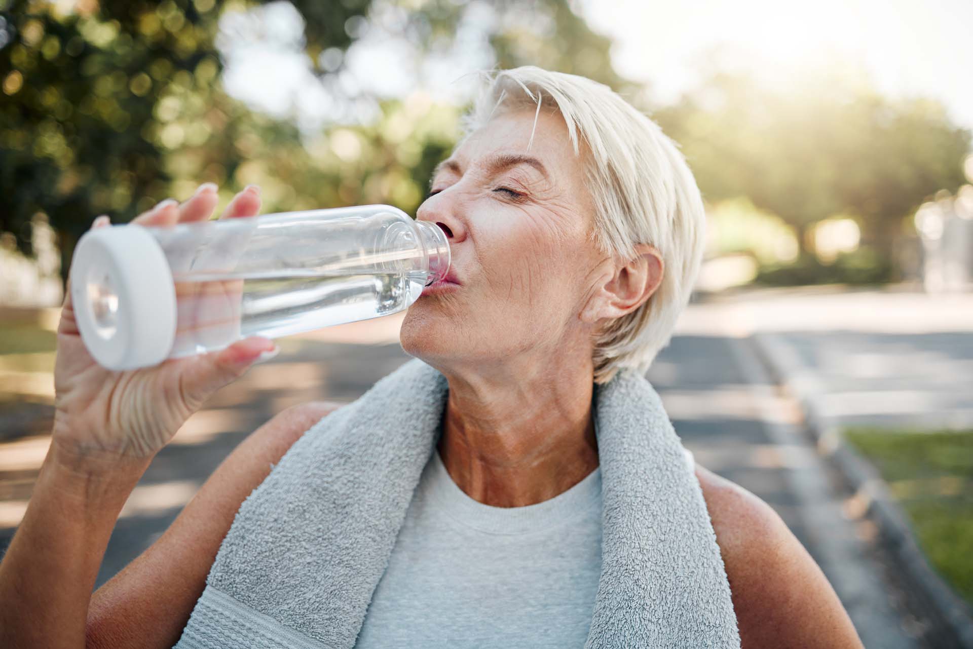 A mature woman with cropped blonde hair drinking from a clear water bottle after outdoor exercise, with a towel around her neck