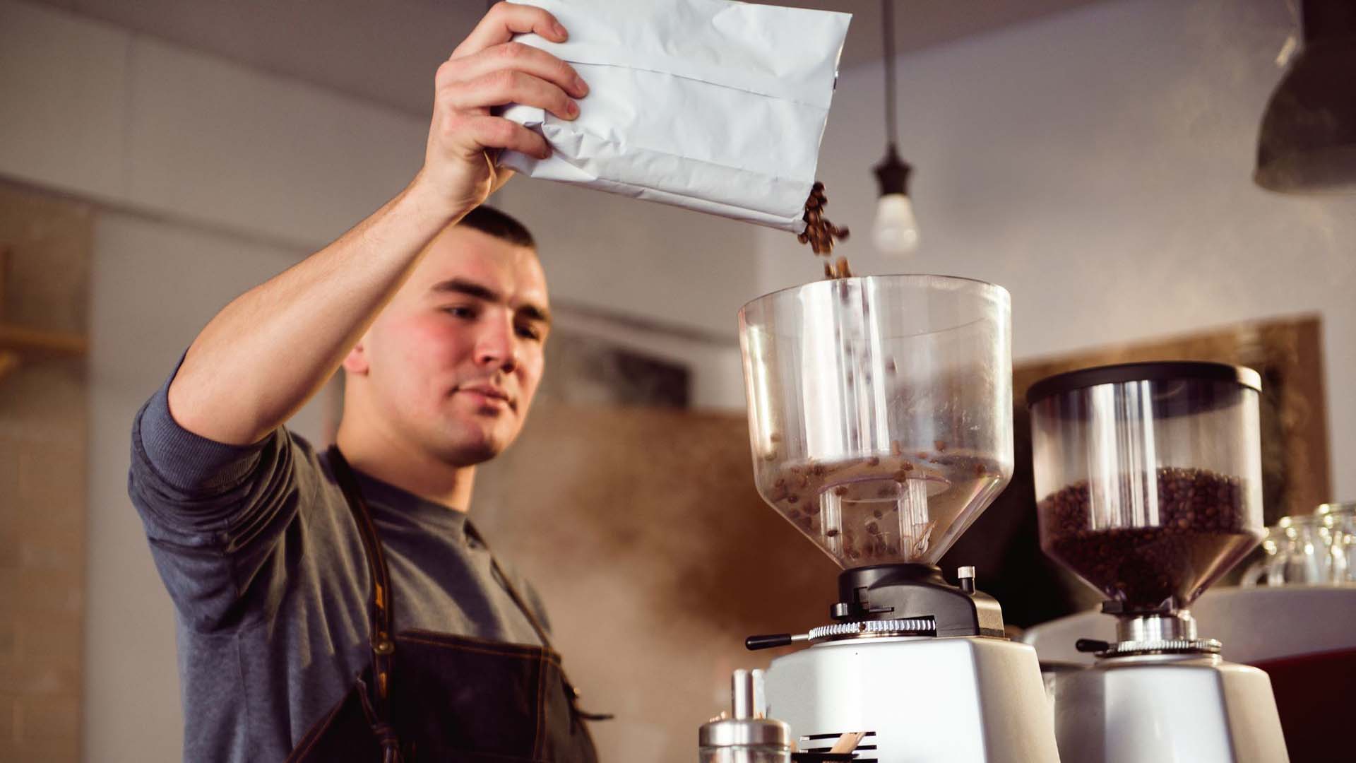 A male barista pours coffee beans into the grinder of a coffee-shop coffee machine
