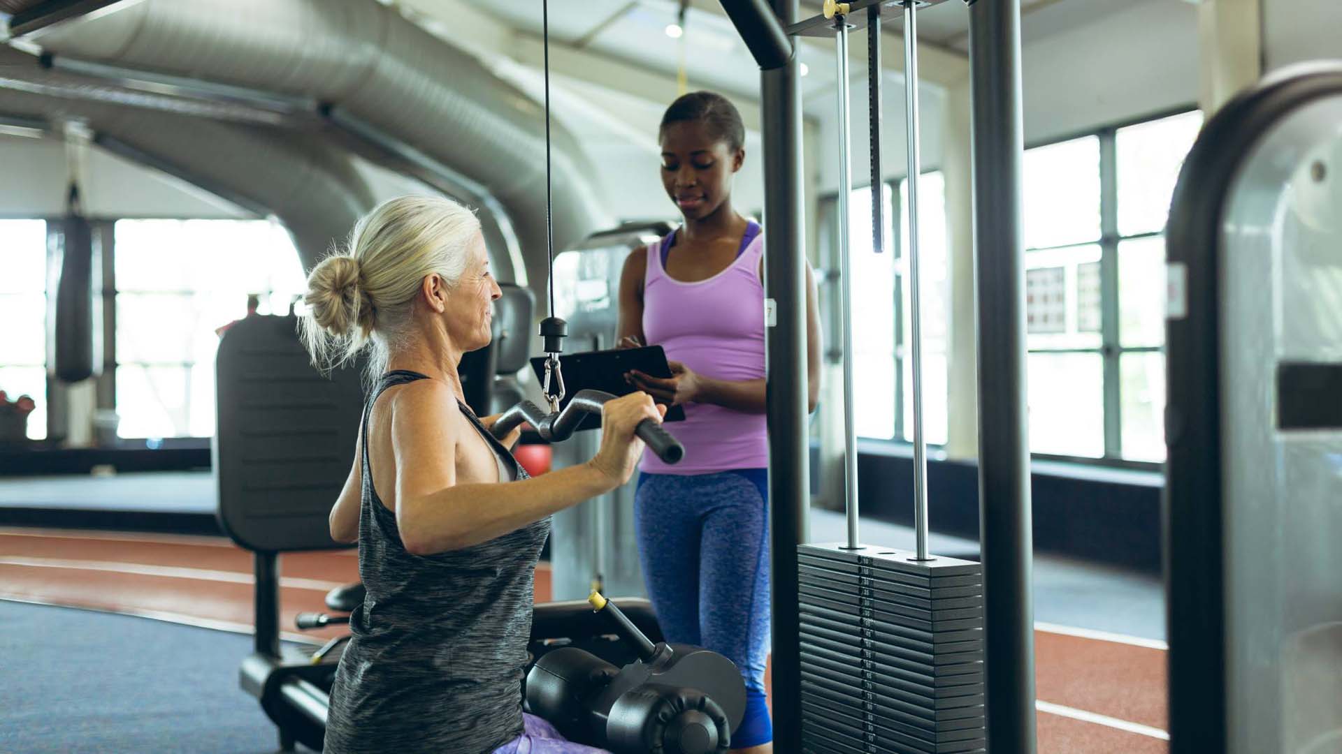 A blonde woman using a weight machine at the gym while a young instructor watches