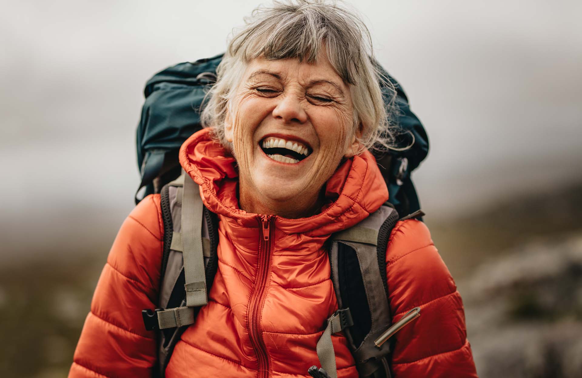 A woman in hiking gear and an orange coat laughs while facing the camera