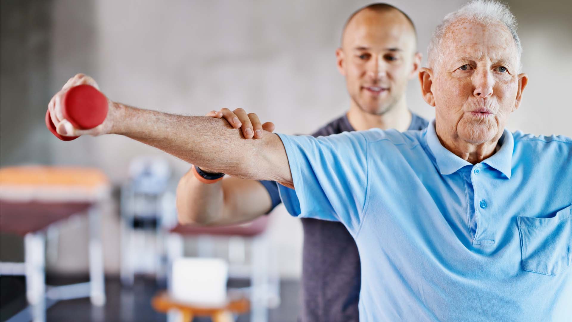 An older man performs side raises with hand weights with an instructor checking his form