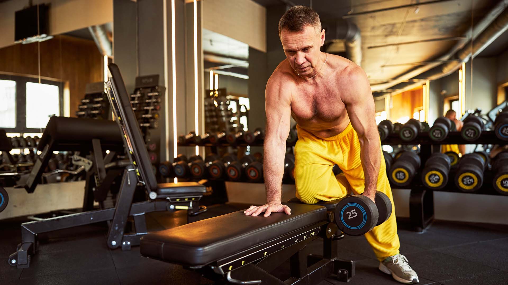 A shirtless man in yellow jogging bottoms lifts a weight while kneeling on a bench in a gym setting