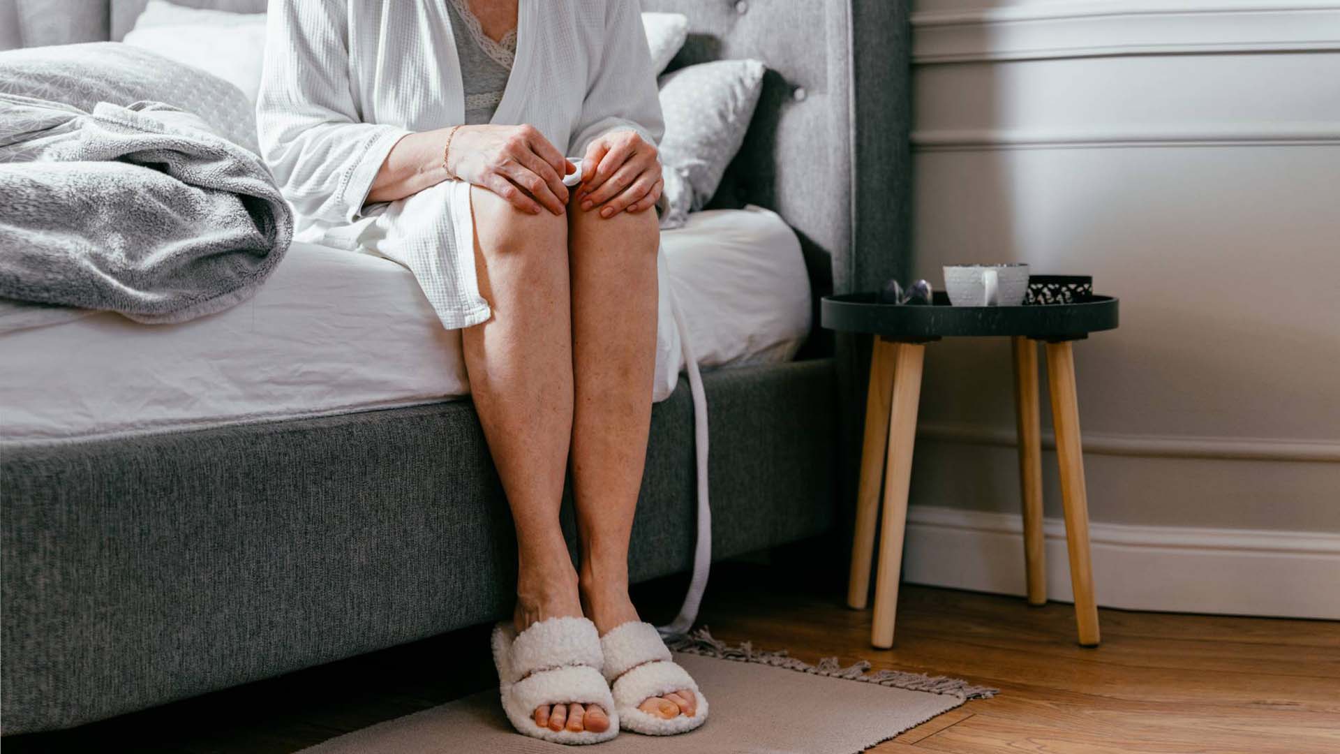 Close up of lonely elderly woman sitting on the bed posing with legs holding knees in arms in front of camera at home against bedroom.