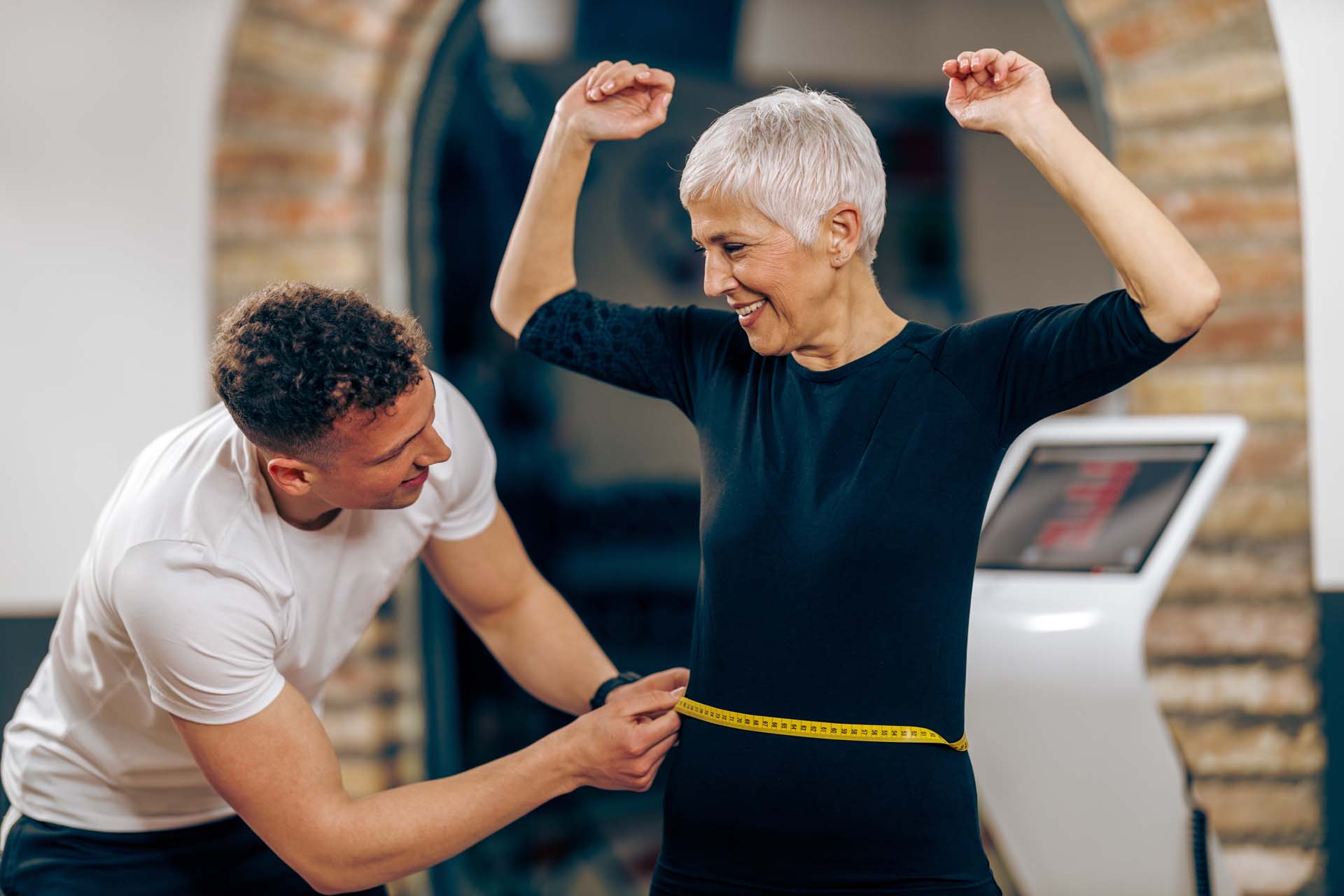 A woman with cropped white hair has her waist measured by a fitness instructor