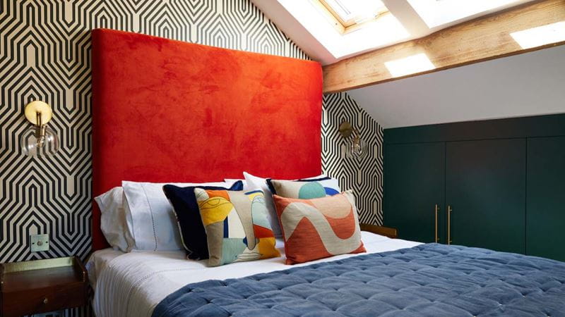 A bed with orange headboard and bright cushions against a geometric black wallpaper in an attic room