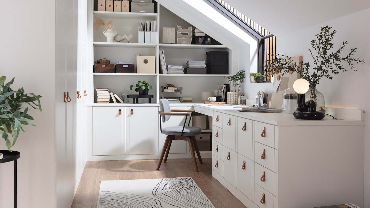 A modern home office with large built-in desk, drawers and wall shelving in white, with black furnishings such as a desk chair and side table