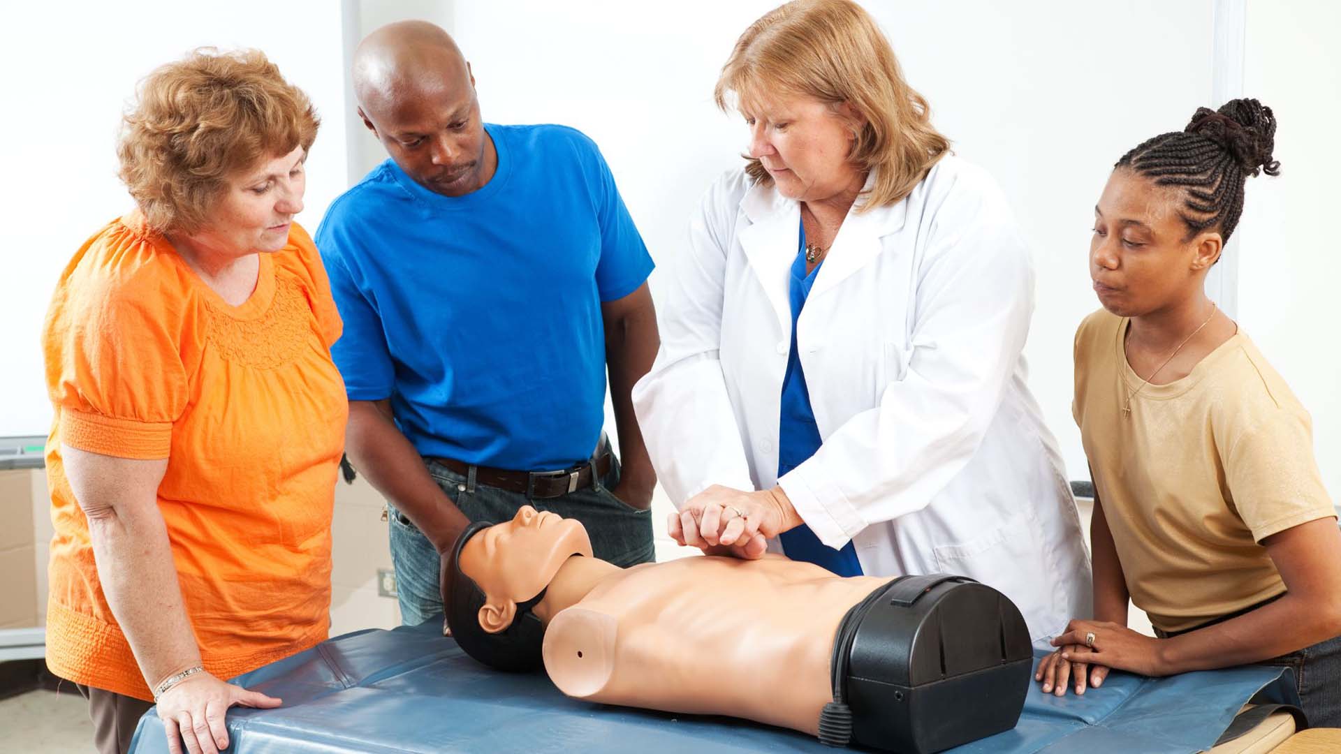 An instructor in a white coat performs CPR on a dummy as part of a first-aid course, with three adults observing