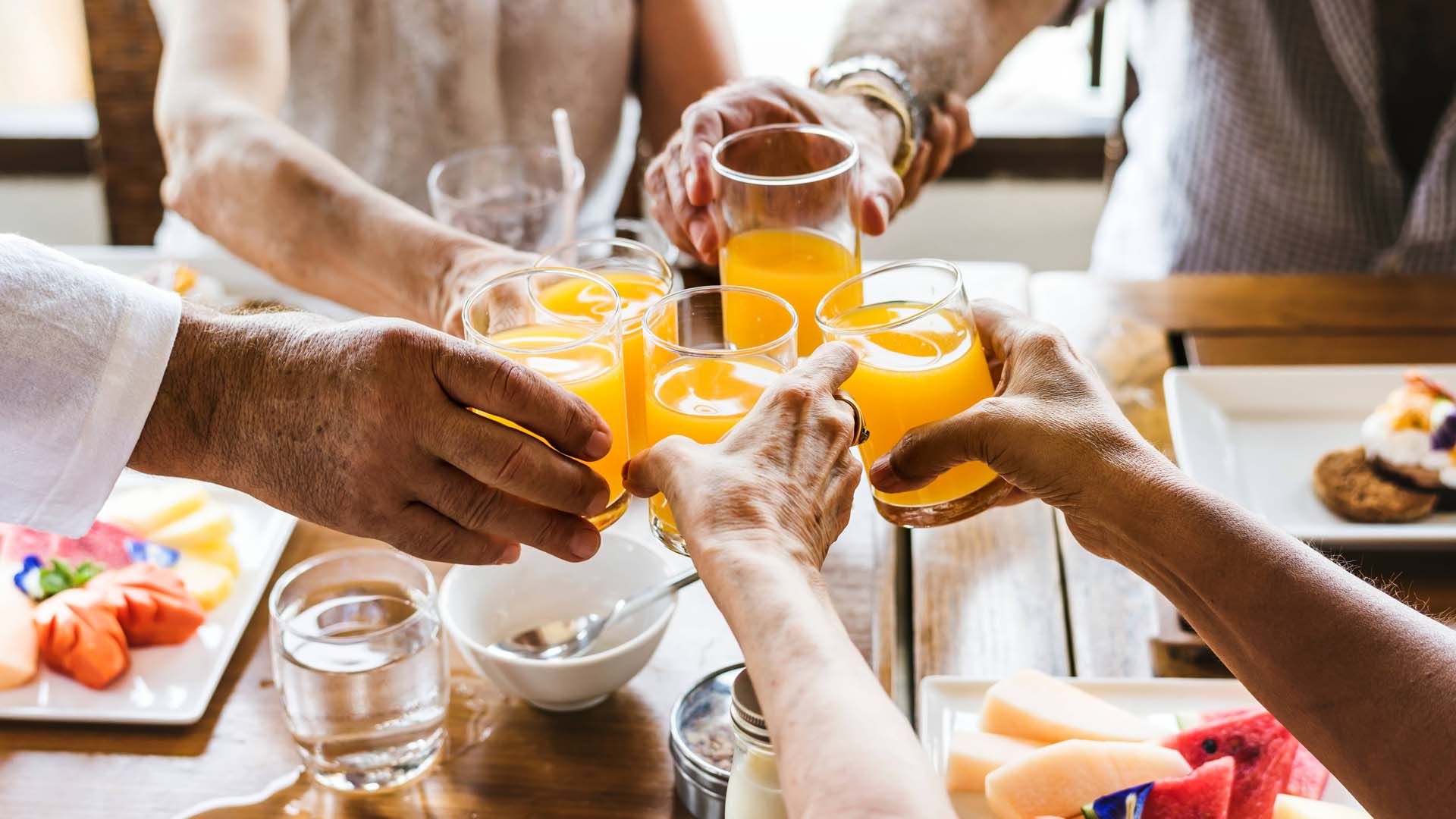 Group of  five people around a table, only their arms showing and clinking orange  juice in the middle of the table.