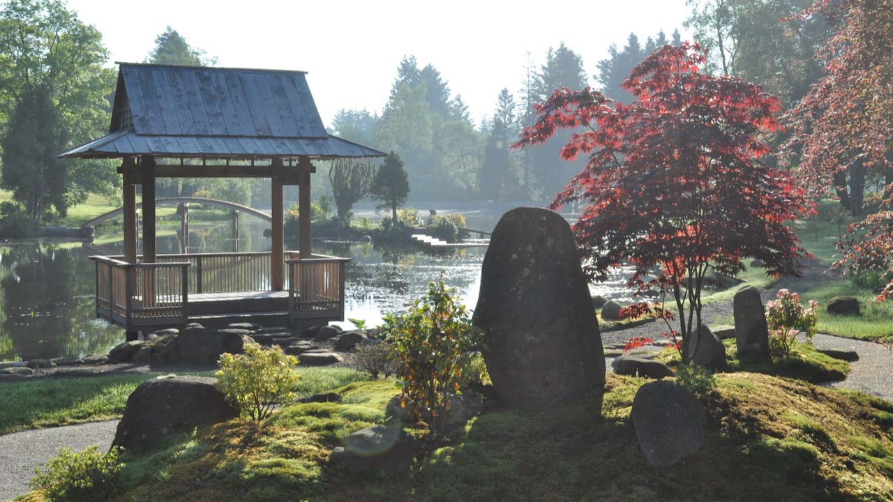 A pergola in a Japanese garden by the water's edge, surrounded by a variety of green and burgundy leaf trees, rocks, and the beautiful hues cast by the setting sun
