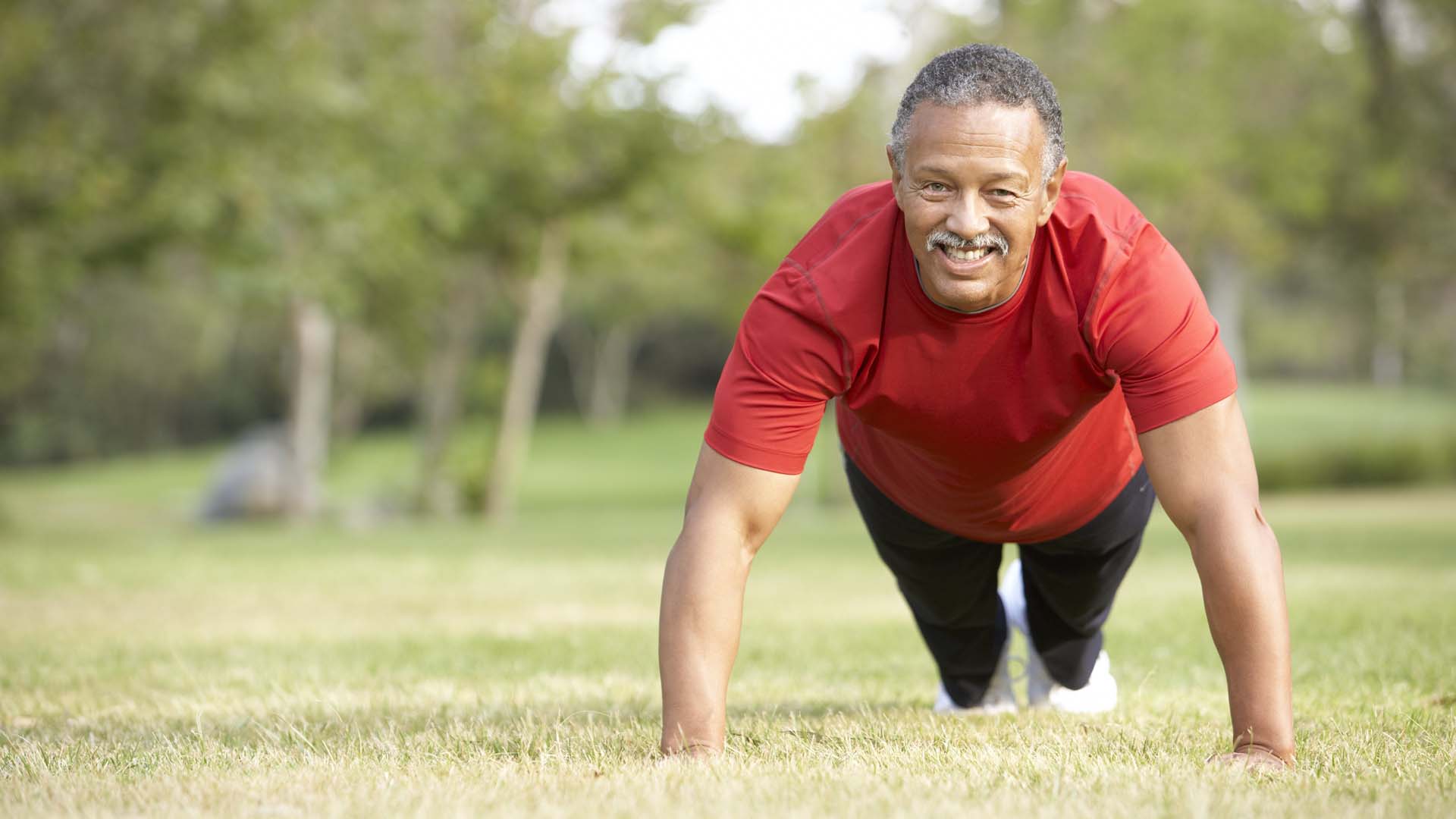 A man in a park in a plank position wearing a red t-shirt
