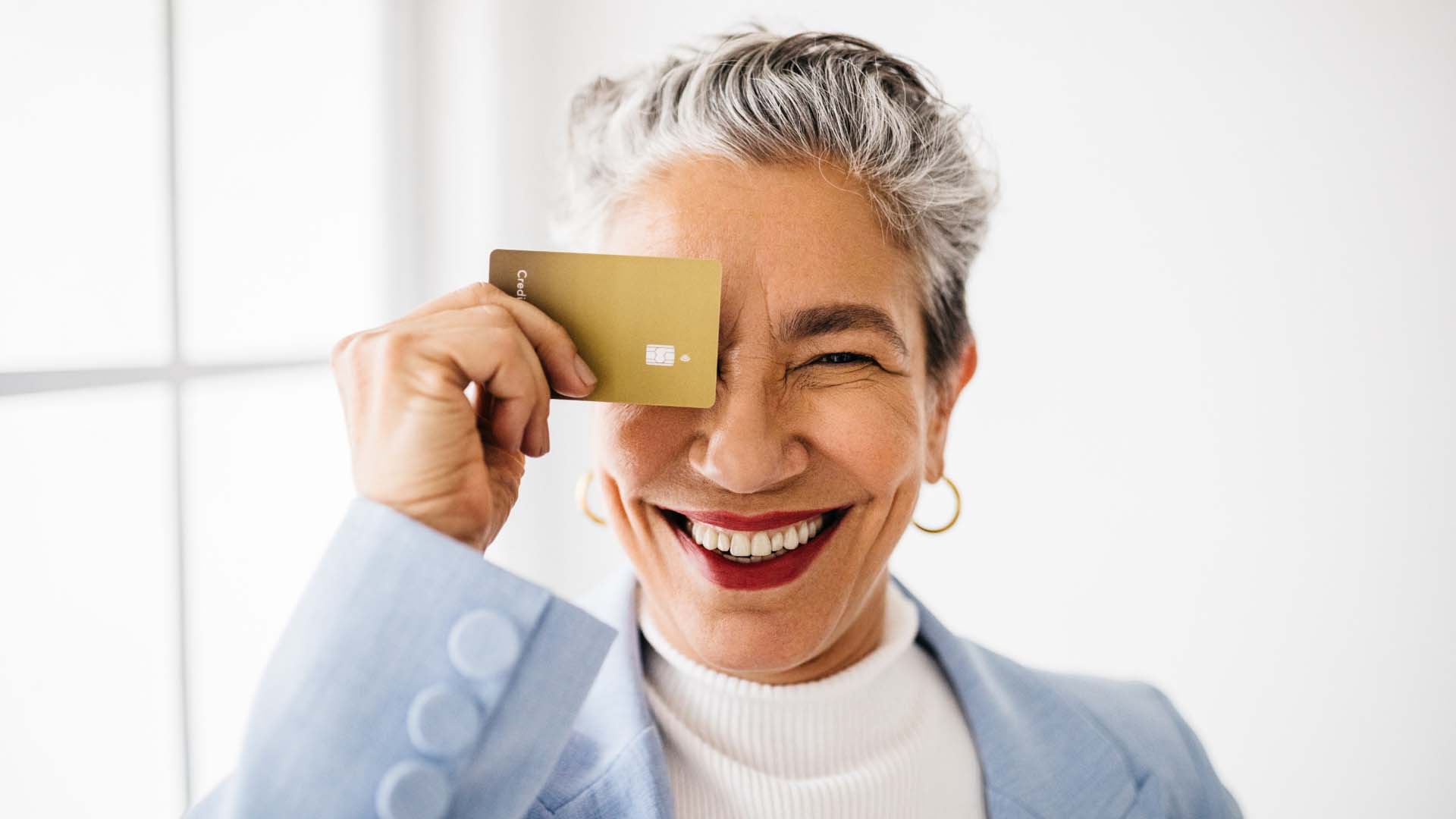 A mature woman in a blue blazer holding up a gold credit card over her eye
