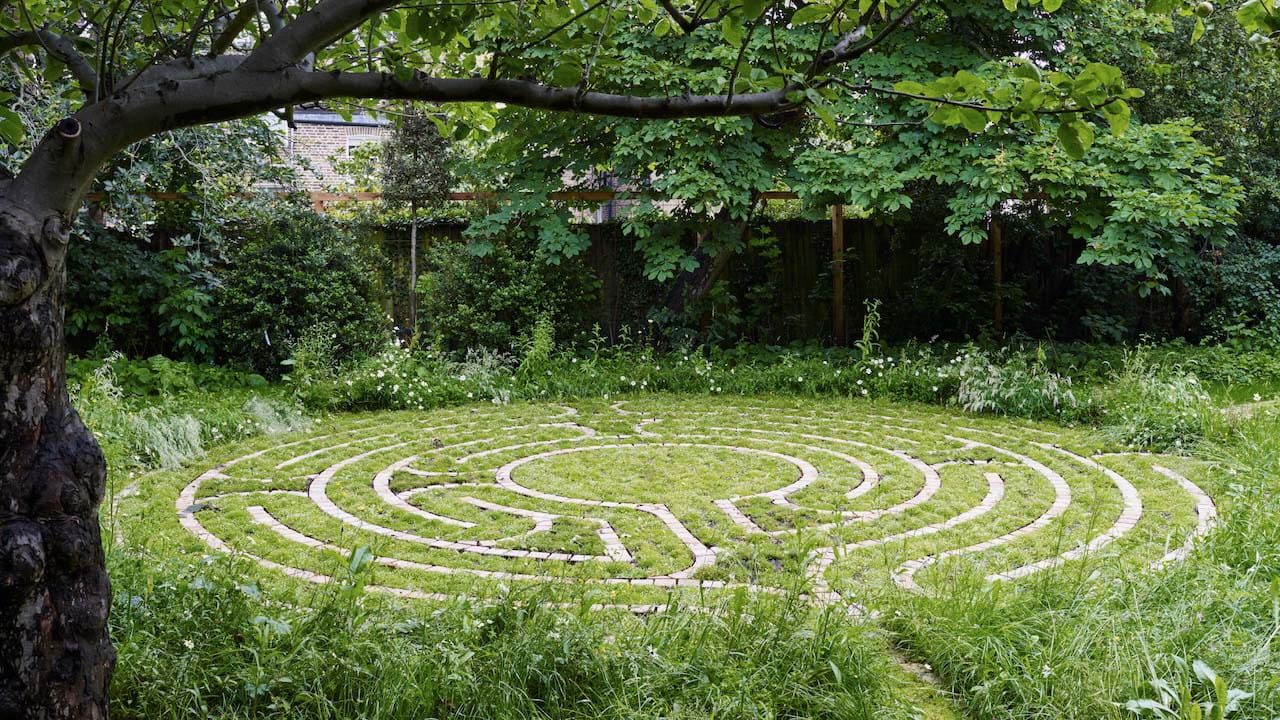 A brick spiral labyrinth embedded in a grassy area beneath the shade of a large, mature tree