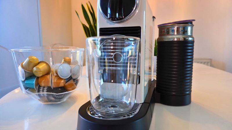 Close-up of a cream coffee pod machine set up with a clear espresso mug, a jar of coffee pods and a milk frother