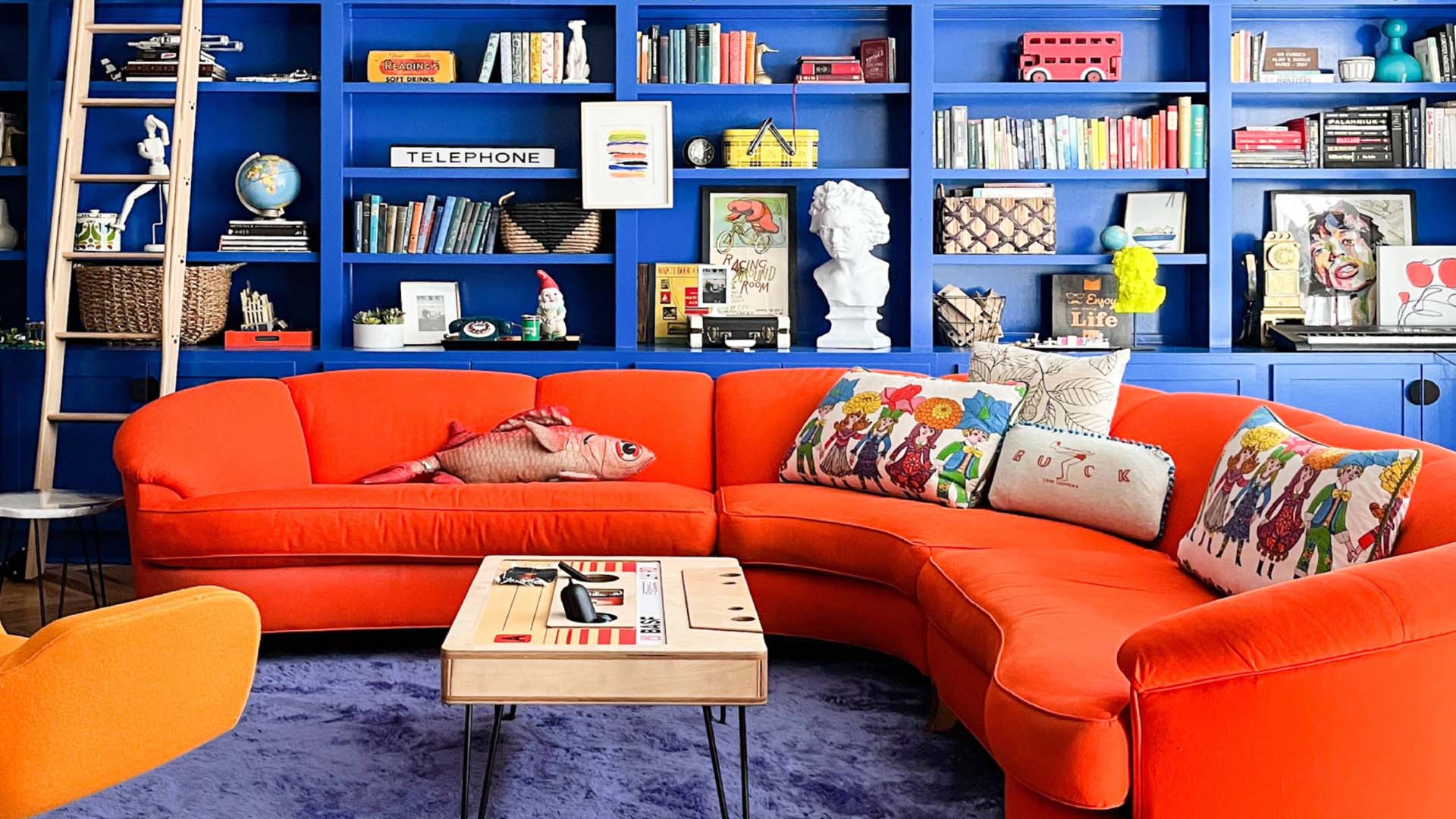 A room with blue bookshelves, a bright orange curved sofa and a blue rug