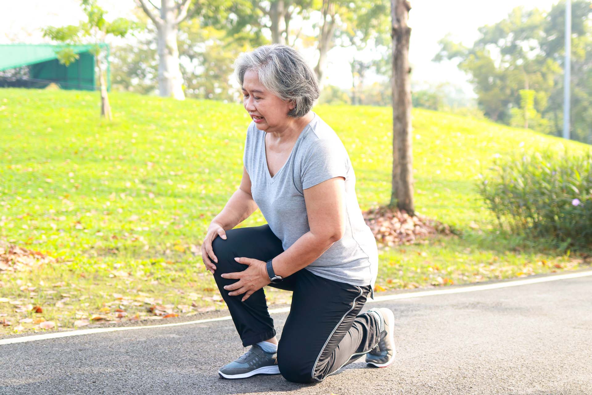 An older woman wearing sports gear kneels to massage a knee joint on a paved path through a park.