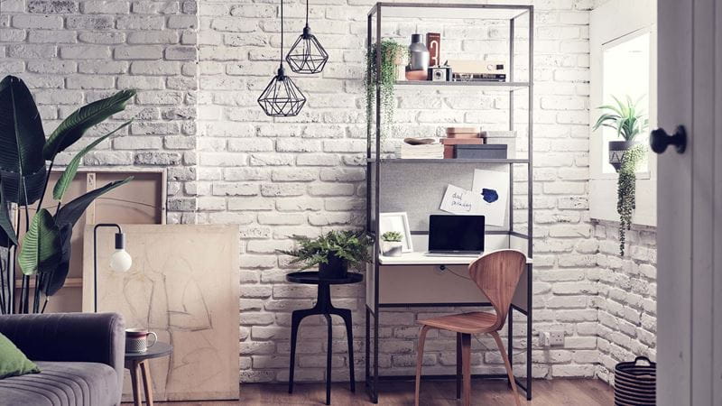 A small home office desk with shelving above against a white brick wall